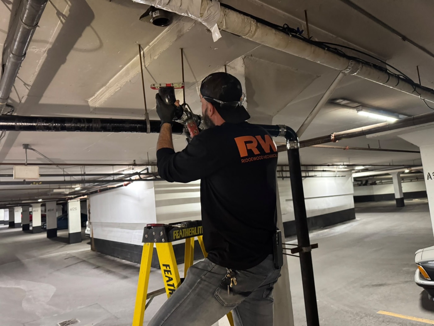 Ridgewood Mechanical technician on a ladder working on overhead piping in a parking garage