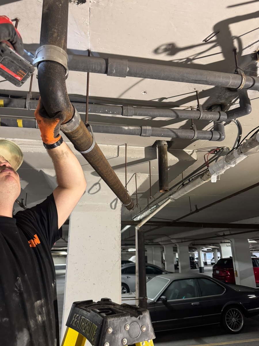 Technician on a ladder working on overhead drain piping in a parking garage